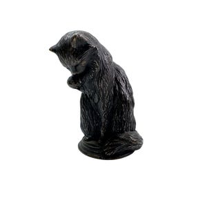 Puede incluir: Una figura de gato en tono bronce oscuro en posición sentada, representada acicalándose la pata. La escultura presenta texturas de pelaje detalladas, orejas puntiagudas y una cola larga envuelta alrededor de su base sobre un pedestal circular.