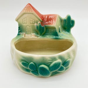 May include: A ceramic planter featuring a house with a red roof and green walls, accented by molded cactus figures. The base is cream-colored with a green relief of prickly pear cactus pads along the front, creating a desert-themed vessel.