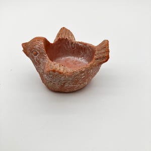 May include: A small, hollow bird-shaped dish crafted from terracotta-colored material with a textured, weathered finish. The piece features a carved head, wings, and tail, with white highlights accentuating the surface details and edges.