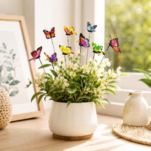 May include: A white ceramic pot containing a cluster of small white flowers and green leaves, accented with several decorative butterfly ornaments on thin wire stems. The butterflies feature vibrant colors including orange, yellow, purple, blue, pink, and green patterns.
