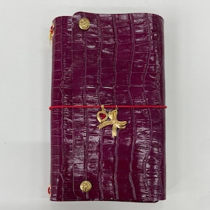 May include: A deep purple, crocodile-embossed leather journal cover featuring two gold-toned circular buttons, a thin red elastic band, and a gold-toned charm set consisting of a small red heart and a decorative bow.