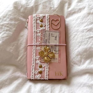 May include: A pink leather journal featuring a vertical white lace trim with gold studs, a gold heart-shaped paperclip, a gold flower charm, and a vintage-style paper insert. The bottom right corner is embossed with the gold letters HK.