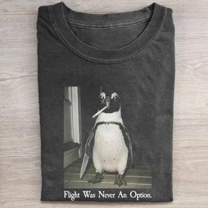 May include: A charcoal gray crew neck t-shirt featuring a graphic of a penguin holding a knife in one flipper and a cigarette in its beak. Below the graphic, the text reads, "Peace Was Never an Option." The shirt has a standard short-sleeve design.