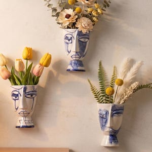 Op de afbeelding: Drie aan de muur gemonteerde keramische vazen in de vorm van menselijke gezichten met blauwe lijntekeningen. Elke vaas bevat een ander arrangement van droogbloemen, waaronder gele craspedia, witte bloemen, eucalyptus en varens.