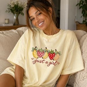 Puede incluir: Una camiseta de manga corta de color amarillo pálido con un estampado gráfico de tres plátanos y dos fresas, cada uno adornado con un pequeño lazo de cuadros verdes. Debajo de la fruta, el texto "just a girl" está escrito en cursiva roja.