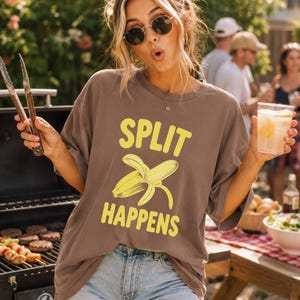 Può includere: Una t-shirt marrone a maniche corte con il testo giallo "SPLIT HAPPENS" sopra un'illustrazione grafica di una banana sbucciata. La maglietta ha uno scollo rotondo e una vestibilità ampia, abbinata a pantaloncini di jeans azzurri.