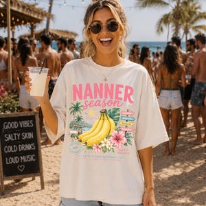Könnte beinhalten: Ein cremefarbenes Oversize-T-Shirt mit einem Grafikdruck mit dem Text NANNER season, einem Bündel gelber Bananen, Palmen, einer rosa Hibiskusblüte und einem kleinen Wegweiser mit verschiedenen Wörtern wie sunshine und banana.