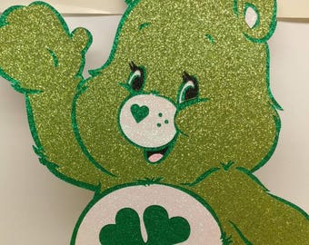 Peut inclure: Ours en peluche vert pailleté avec un trèfle à quatre feuilles sur le ventre. L'ours salue de la patte droite.