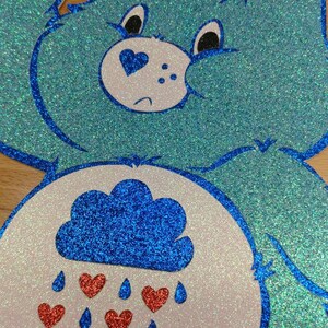 May include: A blue glitter Care Bear with a white belly and a blue cloud with red hearts raining from it.