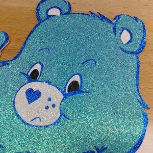 May include: A blue glitter Care Bear with a blue heart on its chest and a white face. The bear has a sad expression with black eyes and a small mouth.