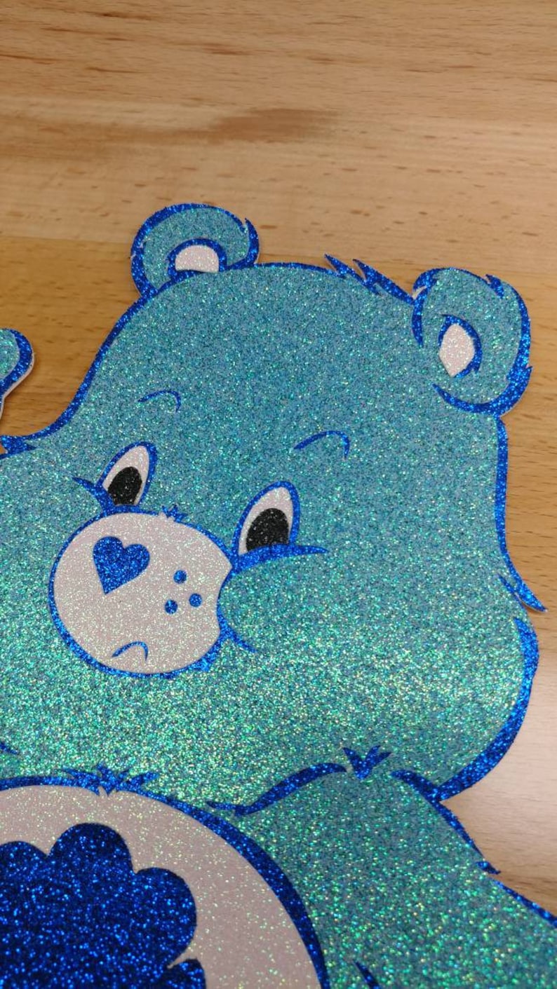 May include: A blue glitter Care Bear with a blue heart on its chest and a white face. The bear has a sad expression with black eyes and a small mouth.