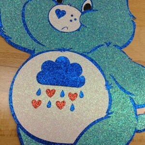 May include: A blue glitter Care Bear with a white belly and a blue cloud with red hearts raining from it. The Care Bear has a sad expression.