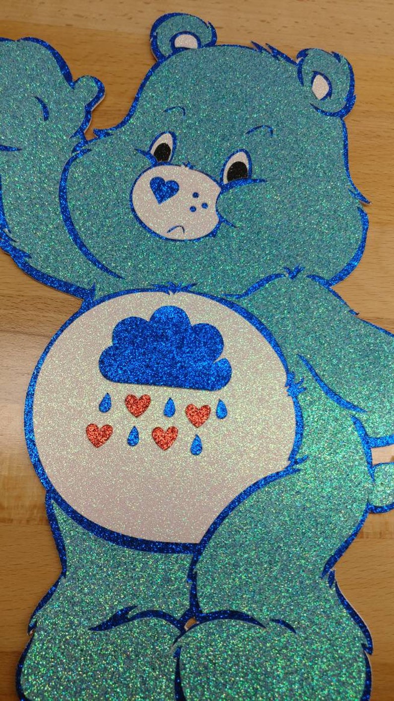 May include: A blue glitter Care Bear with a white belly and a blue cloud with red hearts raining from it. The Care Bear has a sad expression.