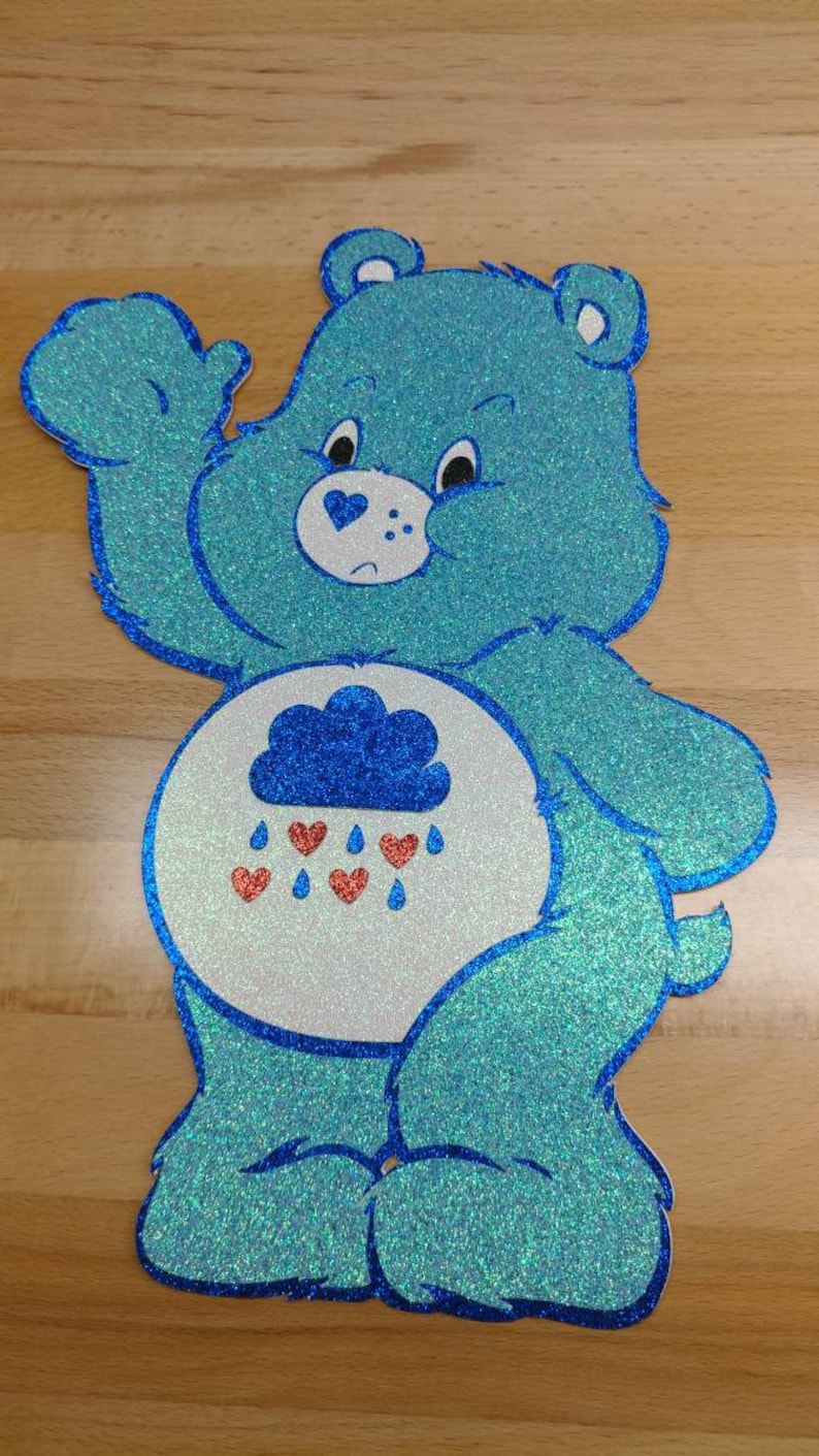 May include: A blue glitter Care Bear with a cloud and raindrops on its belly. The bear has a blue heart on its face and is waving.