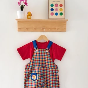 Puede incluir: Una camiseta roja de manga corta bajo un peto con estampado de cuadros amarillos, rojos y azules. El peto tiene tirantes azules con hebillas metálicas y dos bolsillos azules con ribete festoneado amarillo, con un pato y un cangrejo.