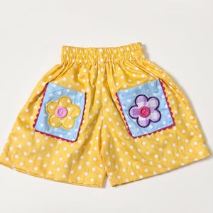 Op de afbeelding: Gele korte broek met een wit stippenpatroon, voorzien van een elastische tailleband en twee lichtblauwe zakken. Elke zak is versierd met een stoffen bloemapplicatie, één geel en één paars geblokt, elk met een roze knoop in het midden.