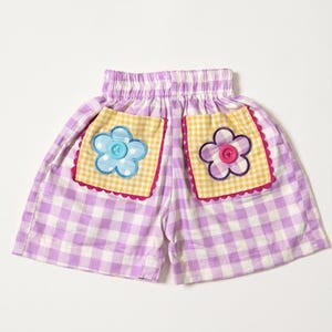 Op de afbeelding: Lila en witte gingham shorts met een elastische tailleband. Twee geel en witte gingham zakken met een geschulpte roze rand, een blauwe bloemapplicatie met een lichtblauwe knoop en een paarse bloemapplicatie met een felroze knoop.