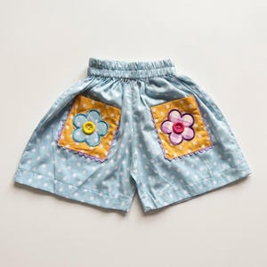 Op de afbeelding: Lichtblauwe shorts met een wit stippenpatroon, voorzien van een elastische tailleband en twee gele vierkante zakken. Elke zak is versierd met een stoffen bloemapplicatie, één lichtblauw en één paars geblokt, met een knoop in het midden.