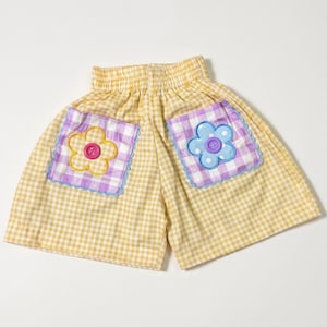 Op de afbeelding: Geel-witte gingham shorts met een elastische tailleband. Twee vierkante zakken met een paars-wit gingham patroon, geschulpte randen en grote bloemvormige stoffen applicaties met een roze knoop links en een paarse knoop aan de rechterkant.