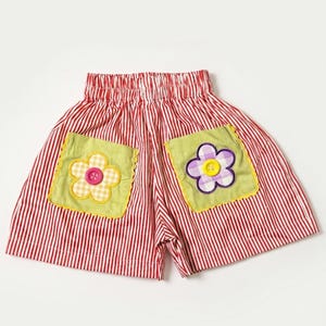 Op de afbeelding: Rood-wit verticaal gestreepte shorts met een elastische tailleband. Aan de voorzijde zijn twee limoengroene vierkante zakken bevestigd, elk voorzien van een stoffen bloemapplicatie met een centrale knoop in respectievelijk roze en geel.