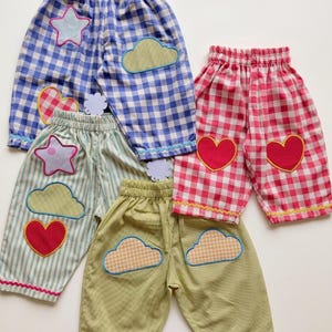 Puede incluir: Una colección de pantalones infantiles en varios patrones y colores. Los pantalones presentan formas aplicadas como estrellas, nubes y corazones. Los diseños incluyen cuadros azules, cuadros rojos, rayas verdes y verde-amarillo liso. Cada par tiene cintura elástica y ribetes decorativos.
