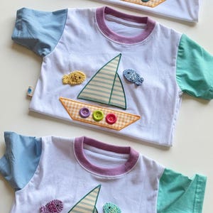 May include: A white t-shirt with a purple collar, light blue left sleeve, and mint green right sleeve. The front features a sailboat applique with a striped sail and a yellow gingham hull decorated with three colorful buttons and two fish.