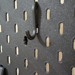 May include: A black pegboard panel featuring a grid of vertical oval-shaped slots and a single black J-shaped hook inserted into one of the openings. The surface of the pegboard has a visible textured finish consistent with 3D printed plastic.