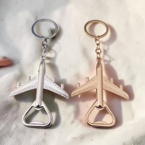 May include: Two metal airplane-shaped bottle openers attached to circular key rings. One bottle opener is silver-toned, and the other is gold-toned. Both feature detailed wings, a fuselage, and a tail section, with the bottle opening mechanism integrated into the rear of the aircraft design.