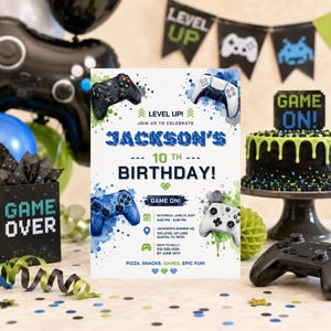 May include: A rectangular birthday invitation card featuring a white background with blue and green watercolor splashes, four game controllers, and text reading Jackson's 10th Birthday, Level Up, Game On, and event details for Saturday, June 21, 2027.