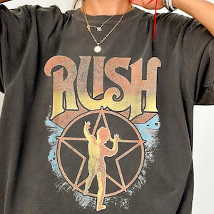May include: Dark gray t-shirt featuring the word "RUSH" in orange and yellow, above a star-shaped design with a figure inside. The shirt has a vintage, worn look.
