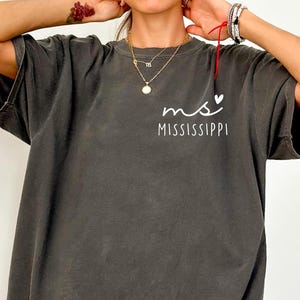 May include: A dark gray t-shirt with the text "ms" above "MISSISSIPPI" in white. The shirt has a relaxed fit and short sleeves. The word "PEPPER" is printed in the bottom right corner.