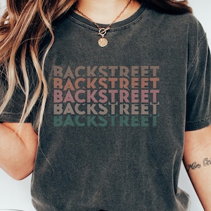 May include: Dark gray t-shirt with the word "BACKSTREET" printed four times in a stacked design. The text colors are brown, pink, mauve, and green. The shirt is a relaxed fit and has a vintage look.