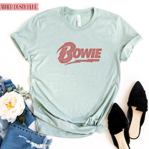 May include: A heather dusty blue t-shirt with a red vintage-style graphic of the word "BOWIE" on the front.
