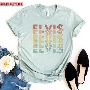 Puede incluir: Una camiseta azul claro con la palabra "ELVIS" repetida en un dise&ntilde;o retro de colores arco&iacute;ris. La camiseta est&aacute; hecha de un material suave y c&oacute;modo.