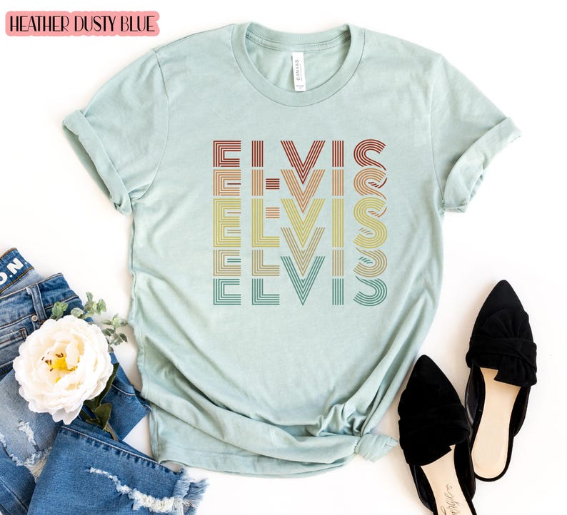 Puede incluir: Una camiseta azul claro con la palabra "ELVIS" repetida en un dise&ntilde;o retro de colores arco&iacute;ris. La camiseta est&aacute; hecha de un material suave y c&oacute;modo.