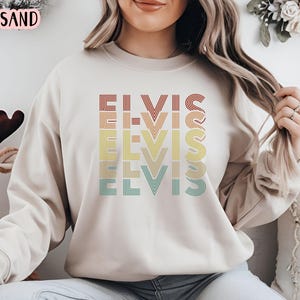 Puede incluir: Una sudadera beige con un gr&aacute;fico de estilo retro de la palabra "ELVIS" repetida en diferentes colores. Los colores del gr&aacute;fico son rojo, naranja, amarillo, verde y azul.