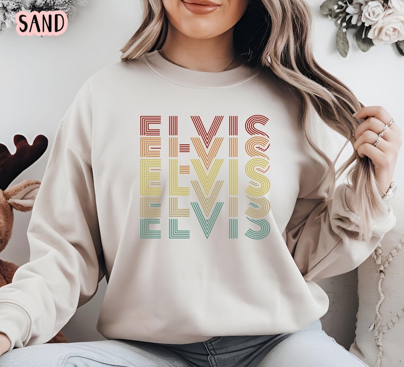 Puede incluir: Una sudadera beige con un gr&aacute;fico de estilo retro de la palabra "ELVIS" repetida en diferentes colores. Los colores del gr&aacute;fico son rojo, naranja, amarillo, verde y azul.