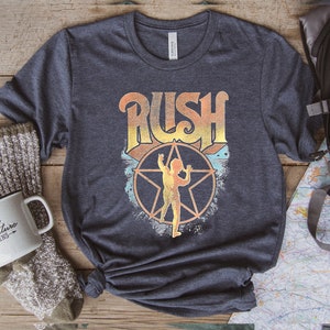 May include: A navy blue t-shirt featuring a vintage-style graphic of the band Rush. The graphic includes the band's name in a stylised font with a star and a figure in the centre.