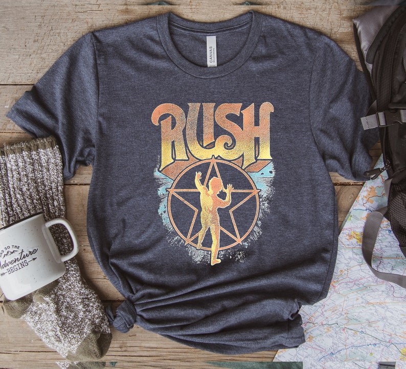 May include: A navy blue t-shirt featuring a vintage-style graphic of the band Rush. The graphic includes the band's name in a stylised font with a star and a figure in the centre.