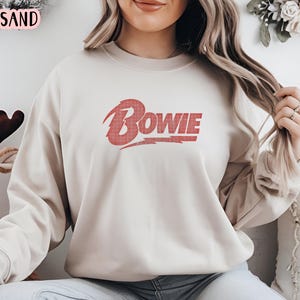 May include: A beige sweatshirt with a red "BOWIE" graphic on the front. The graphic is in a vintage style with a lightning bolt.