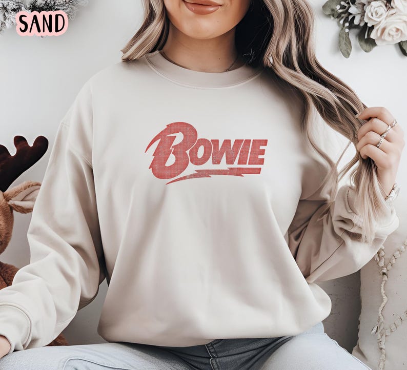 May include: A beige sweatshirt with a red "BOWIE" graphic on the front. The graphic is in a vintage style with a lightning bolt.