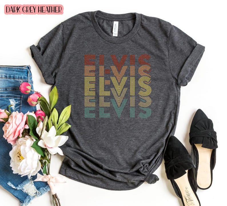 May include: A dark gray heather t-shirt with the word 'ELVIS' printed on it in a retro style with colorful stripes. The word is repeated four times, stacked vertically, with each repetition in a different color. The t-shirt is displayed on a white background with a pair of black shoes and a pair of blue jeans.