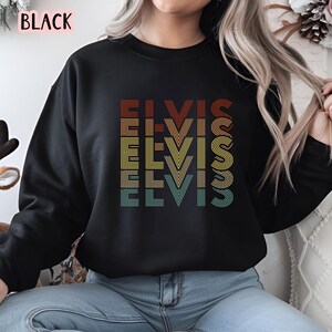 Puede incluir: Sudadera negra con un gr&aacute;fico de estilo retro de la palabra "Elvis" repetida en diferentes colores y fuentes.