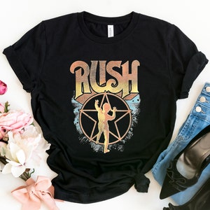 May include: Black t-shirt featuring a vintage-style graphic of the band Rush. The graphic includes the band's name in a stylised font with a star and a figure in the centre.