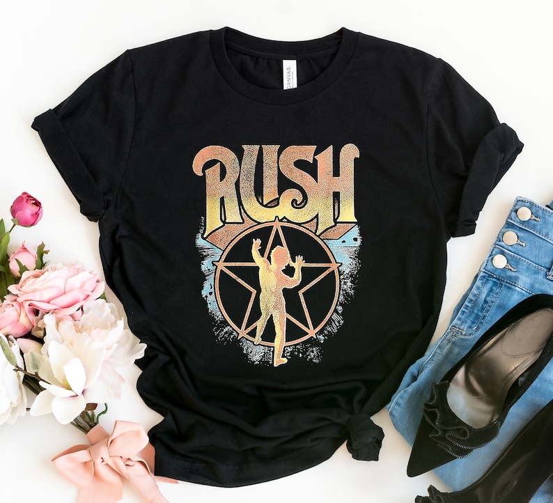 May include: Black t-shirt featuring a vintage-style graphic of the band Rush. The graphic includes the band's name in a stylised font with a star and a figure in the centre.