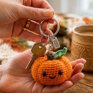 May include: A crocheted orange pumpkin keychain with a brown stem, a green leaf, and two black bead eyes. The keychain features a silver metal ring, a silver key, and a small rectangular silver tag with the text "PURRAIN I FALL FOR WITCHIN".