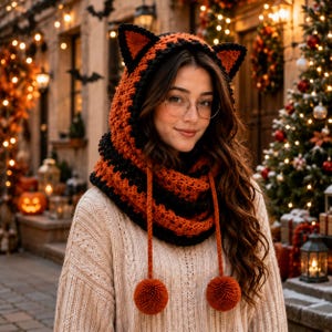 Peut inclure: Une capuche au crochet avec des oreilles de chat dans un motif rayé orange et noir. La capuche comprend deux longs cordons pendants, chacun terminé par un pompon orange rond. Le design est réalisé avec un point de tricot texturé.