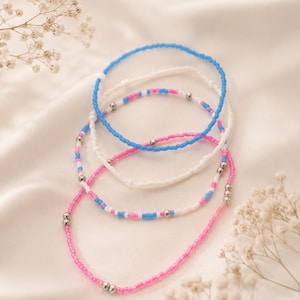 May include: Four beaded bracelets in shades of pink, white, and blue. The bracelets are made of small, colorful beads strung together with silver-colored metal accents. The bracelets are displayed on a white, silky surface.