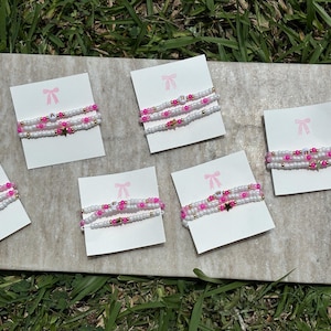 May include: Five sets of three beaded bracelets displayed on white cards with a pink bow icon. Each bracelet features white and pink beads, small gold-tone spacers, and occasional star-shaped charms or letter beads, arranged in a stack of three.