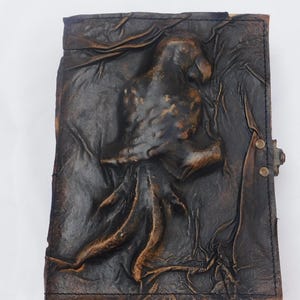 May include: A dark brown leather journal featuring a raised, embossed parrot design on the front cover. The leather has a distressed, textured finish with visible stitching along the edges and a small metal latch closure on the right side.
